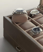 Three luxury watches are elegantly displayed in a TAWBURY Bayswater Watch Box, with one resting on an elevated cushion. Slender beige velveteen wrist pillows (50mm) cradle each timepiece, adding sophistication to the brown leather setting.